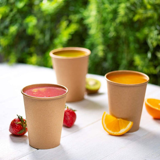 Three paper cups with colorful drinks on a table outdoors, surrounded by fruits and greenery.